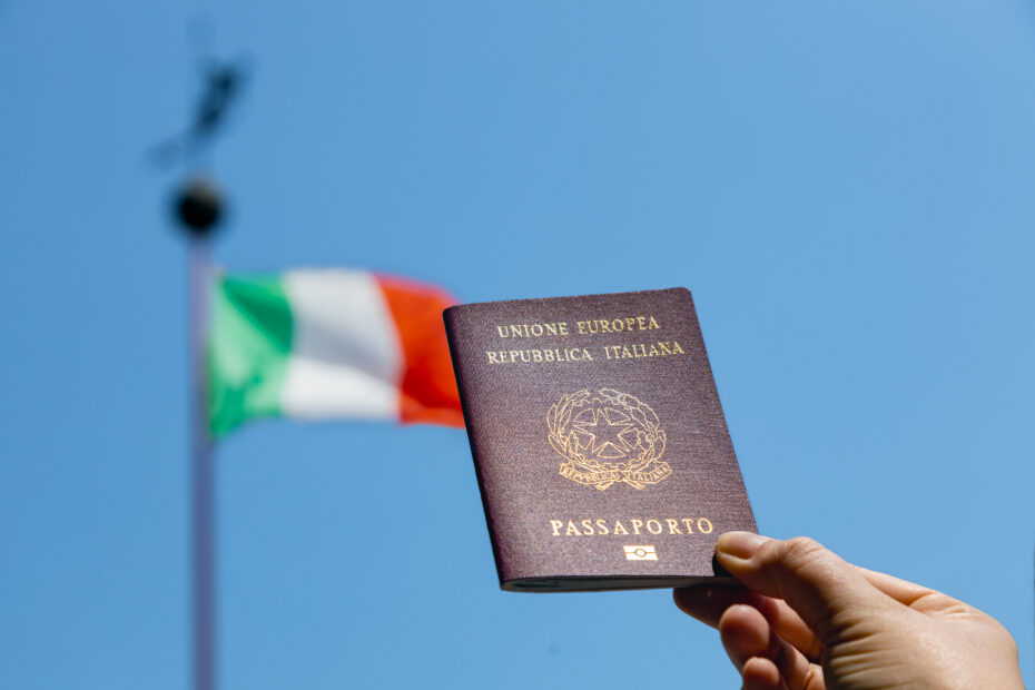 Imagem da bandeira italina sob o fundo azul do céu e uma mão segurando um passaporte da Itália.